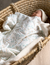 Neugeborenes Baby liegt in einem Moseskörbchen und wird von einer Babydecke mit kleinen Weidenzweigen in zarten Pastelltönen mint und helles braun zudedeckt